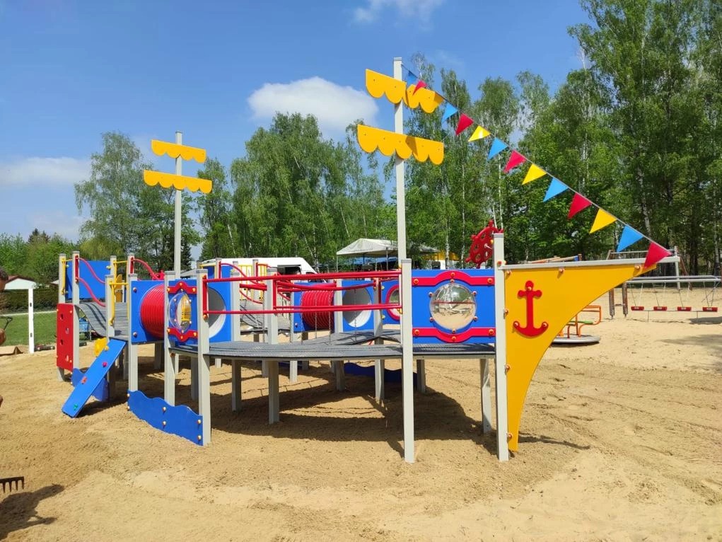 Kolorowy plac zabaw w kształcie statku na piaszczystym terenie, z drzewami w tle i elementami do wspinaczki, zjeżdżania i zabawy.
