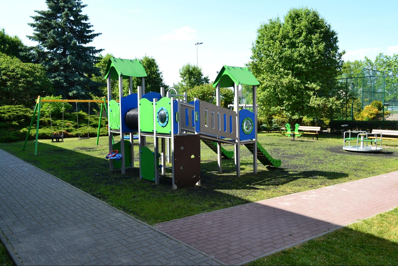 Kolorowy plac zabaw z huśtawką, zjeżdżalnią i karuzelą na trawiastym terenie, otoczony zielenią. Widok na plac zabaw z perspektywy chodnika.