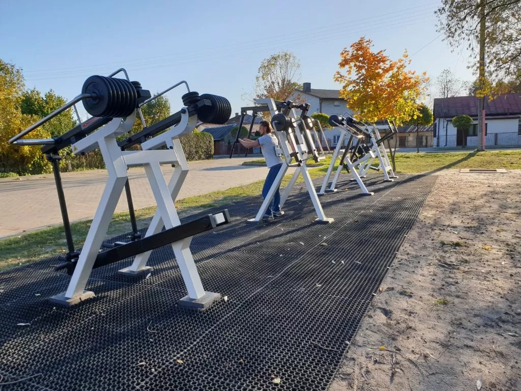 Zestaw urządzeń do ćwiczeń na świeżym powietrzu z obciążeniami, ustawionych rzędowo na czarnej, gumowej nawierzchni, z osobą ćwiczącą na jednym z nich.