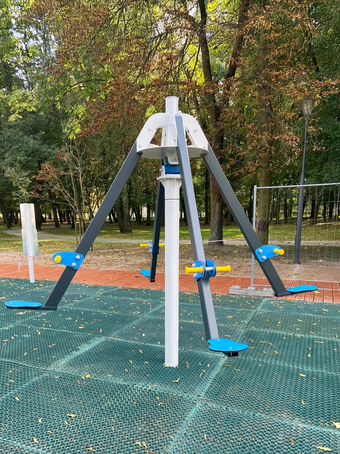 Atrakcja na placu zabaw: karuzela z czterema stanowiskami do ćwiczeń, w kolorach szarym, białym, niebieskim i żółtym, na tle parku.