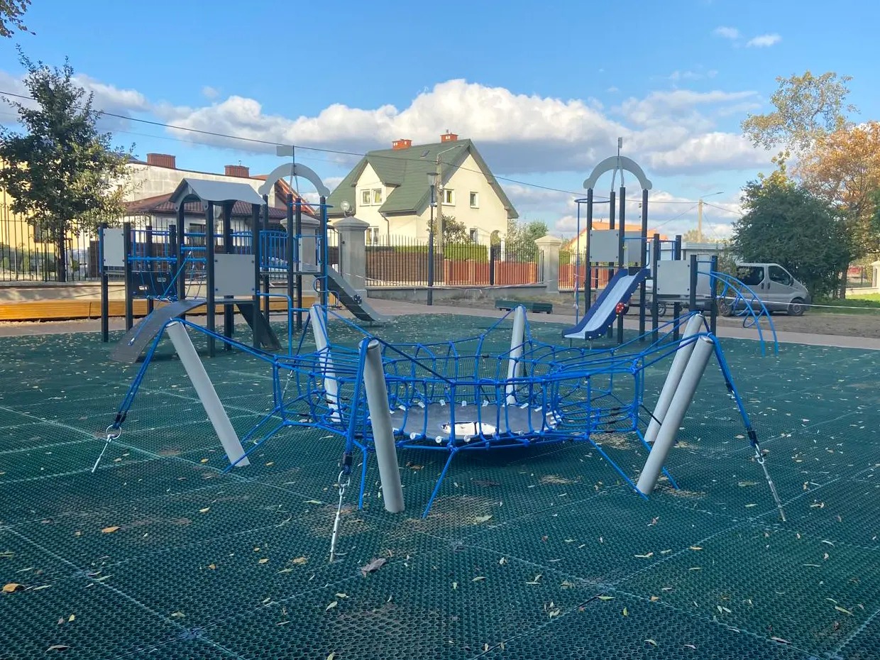 Nowoczesny plac zabaw z siatką pająka, zjeżdżalniami i elementami w kolorze niebieskim, na tle zielonej nawierzchni i zabudowy mieszkalnej.