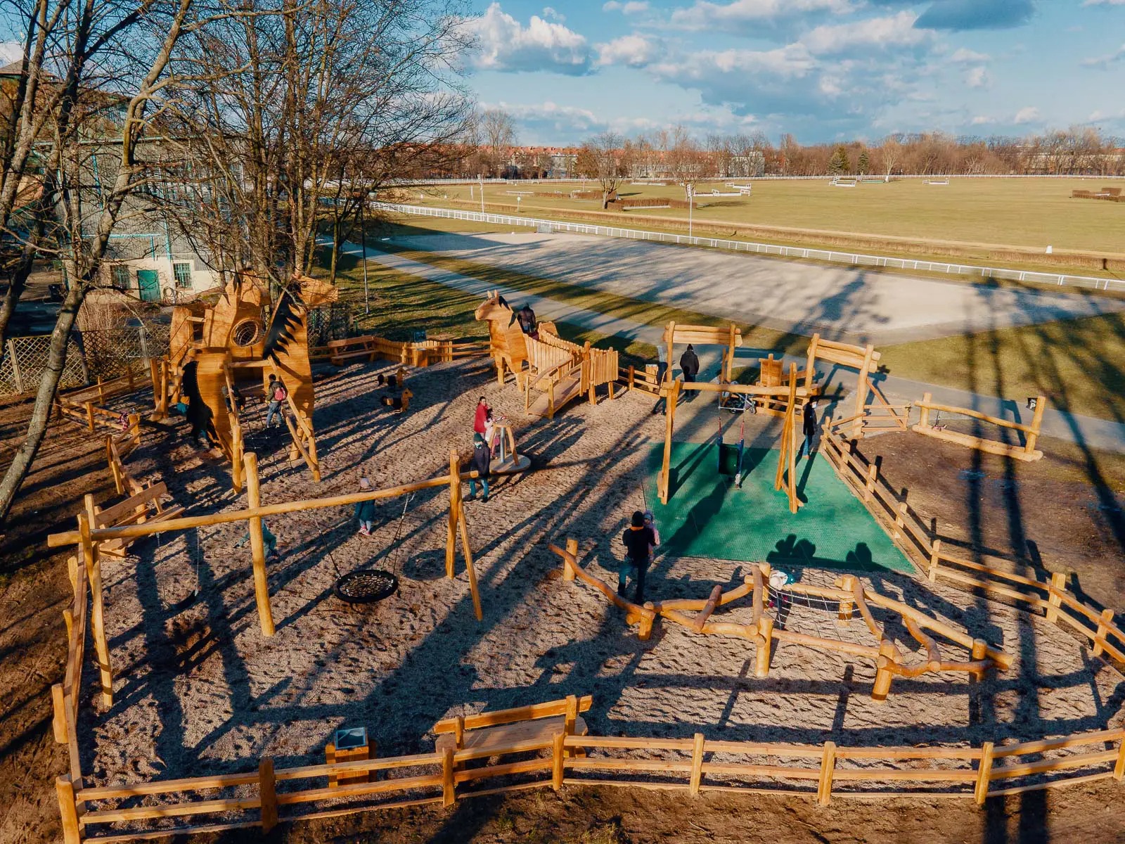 Rozległy plac zabaw z drewnianymi konstrukcjami koni, huśtawkami i piaskiem, otoczony niskim płotkiem. W tle widoczne tereny zielone i budynki.