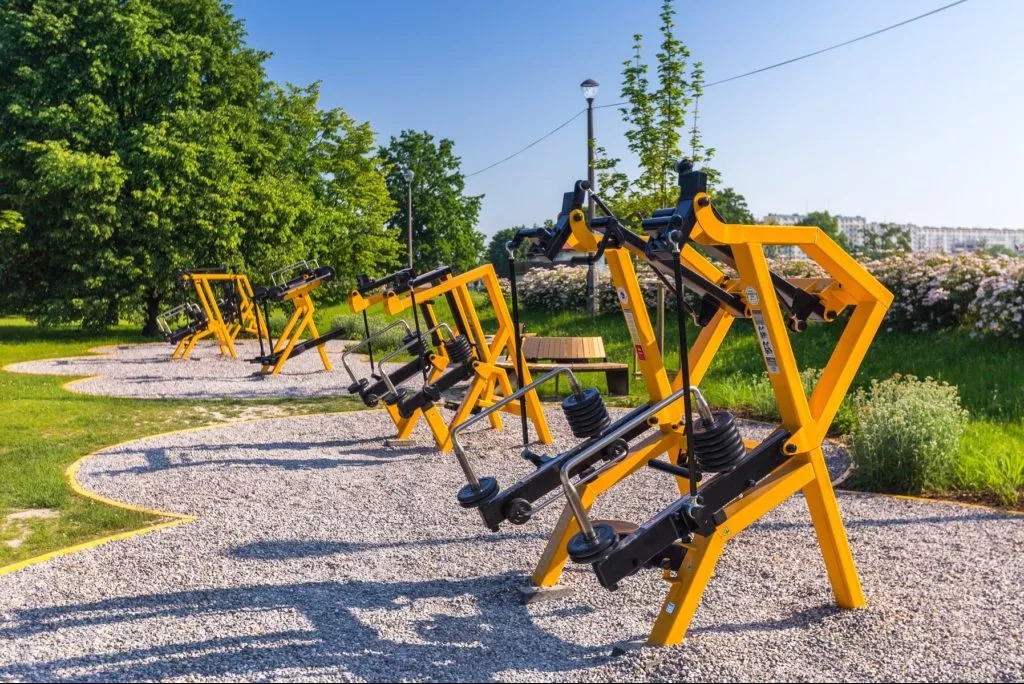 Żółte urządzenia do ćwiczeń na świeżym powietrzu, ustawione w rzędzie na żwirowym podłożu, otoczone zielenią parku i kwitnącymi krzewami.