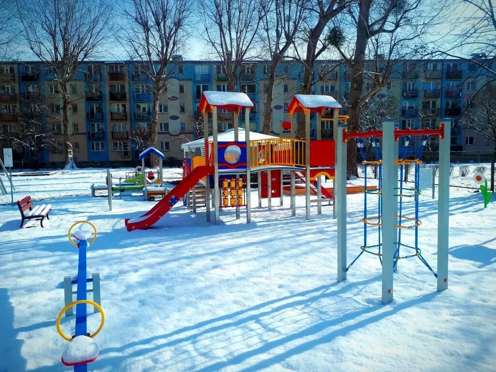 Zimowy plac zabaw z kolorowymi urządzeniami rekreacyjnymi pod blokiem. Śnieg pokrywa teren, rzucając długie cienie. Widoczne drzewa bez liści i niebieskie niebo.