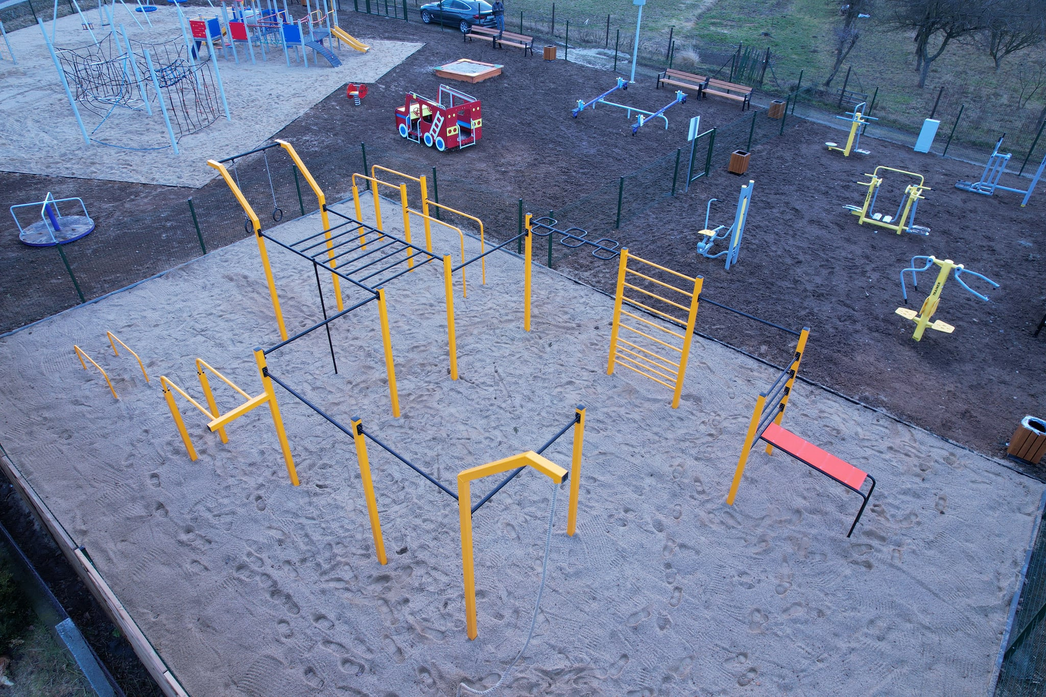 Widok z góry na piaszczysty plac zabaw z żółto-czarnymi elementami do ćwiczeń, huśtawkami i karuzelą w tle. Częściowo ogrodzony, z widocznym samochodem.