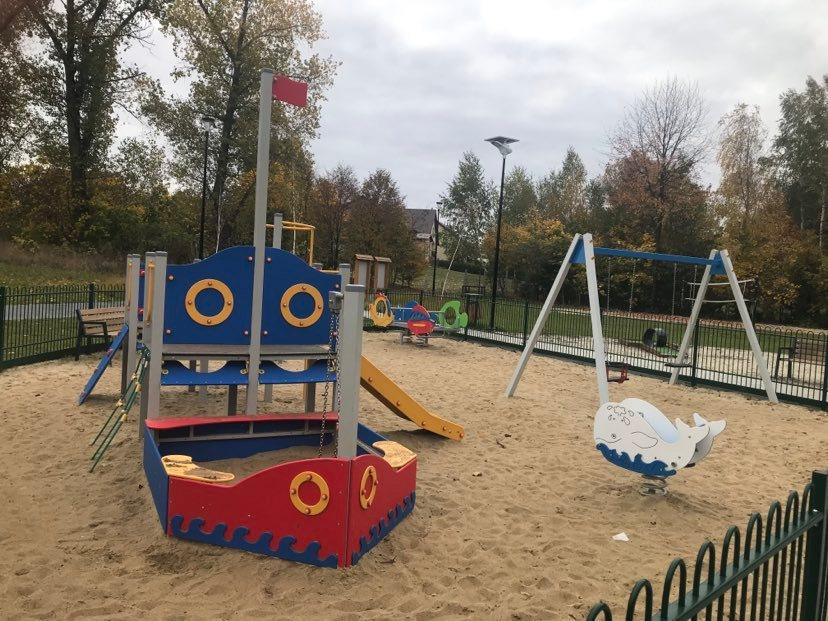 Kolorowy plac zabaw z piaskownicą w kształcie statku, huśtawką ze sprężyną w kształcie wieloryba i klasycznymi huśtawkami na tle jesiennego krajobrazu.