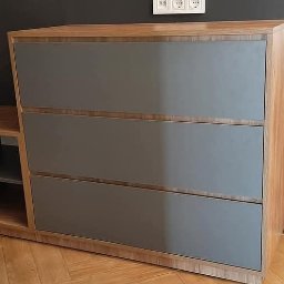 Craftys Mykhailo Tysiachnyi - Drewniana komoda z szarymi frontami szuflad i półkami obok, na tle ciemnej ściany i parkietu. Widoczne buty i fragment stołu w tle.