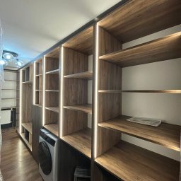 Craftys Mykhailo Tysiachnyi - Wnętrze z zabudową drewnianą z podświetleniem LED, integrującą pralkę. Widoczne półki i schowki, tworzące funkcjonalną przestrzeń. Ujęcie z perspektywy podłogi.