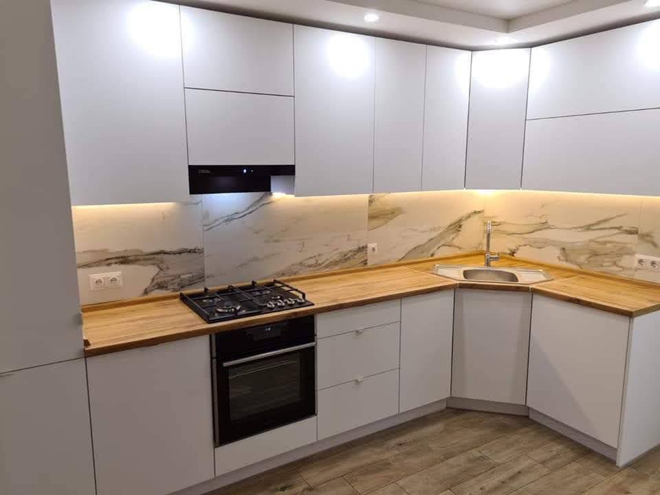 Biała kuchnia z drewnianym blatem i marmurowym panelem. Uwagę przykuwa narożny zlew i oświetlenie LED pod szafkami, które podkreśla wzór marmuru.