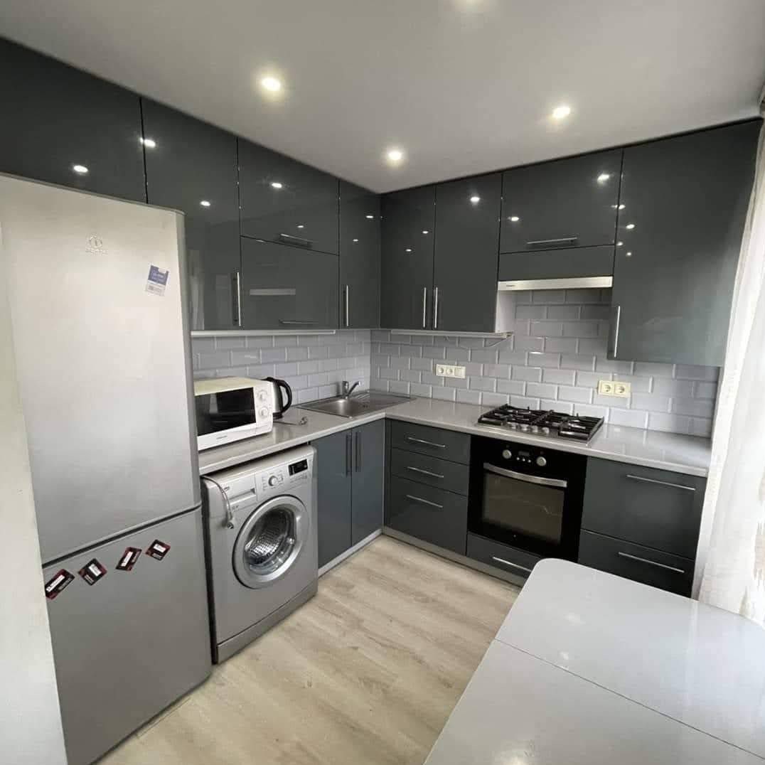 Szara kuchnia z połyskiem, zabudowa narożna z AGD: lodówka, pralka, piekarnik. Białe płytki na ścianie i drewnopodobna podłoga. Nowoczesne oświetlenie.