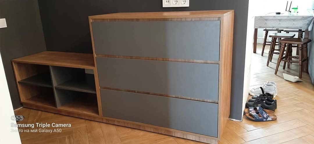 Drewniana komoda z szarymi frontami szuflad i półkami obok, na tle ciemnej ściany i parkietu. Widoczne buty i fragment stołu w tle.