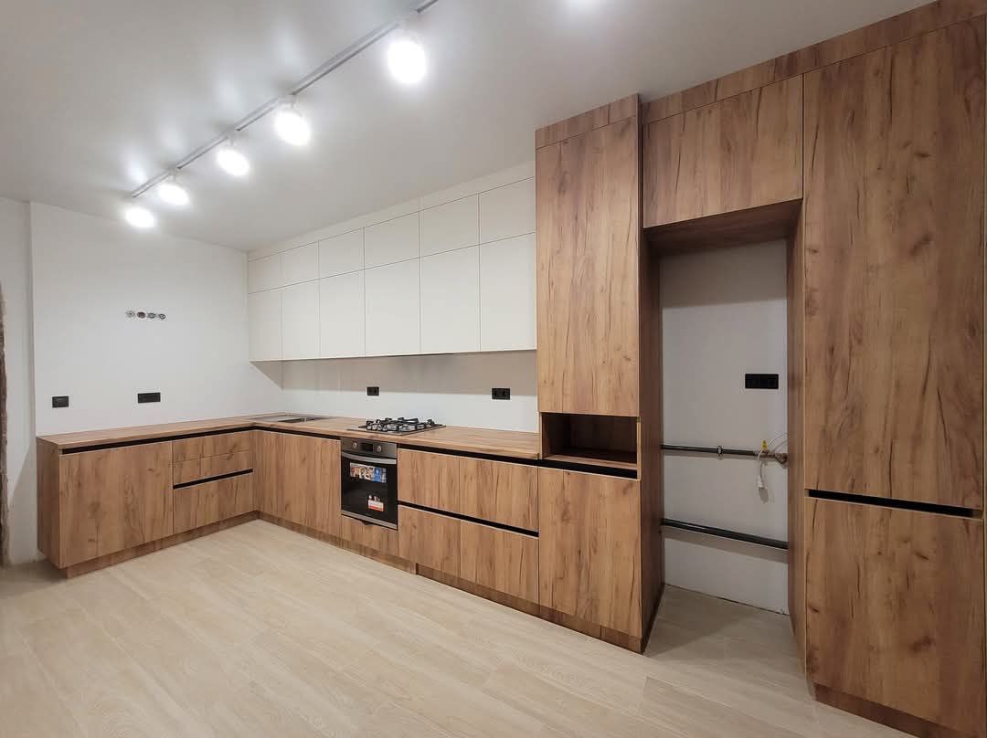 Nowoczesna kuchnia z drewnianymi frontami dolnych szafek i białymi górnymi, oświetlona reflektorami. Widoczny piekarnik i płyta gazowa.