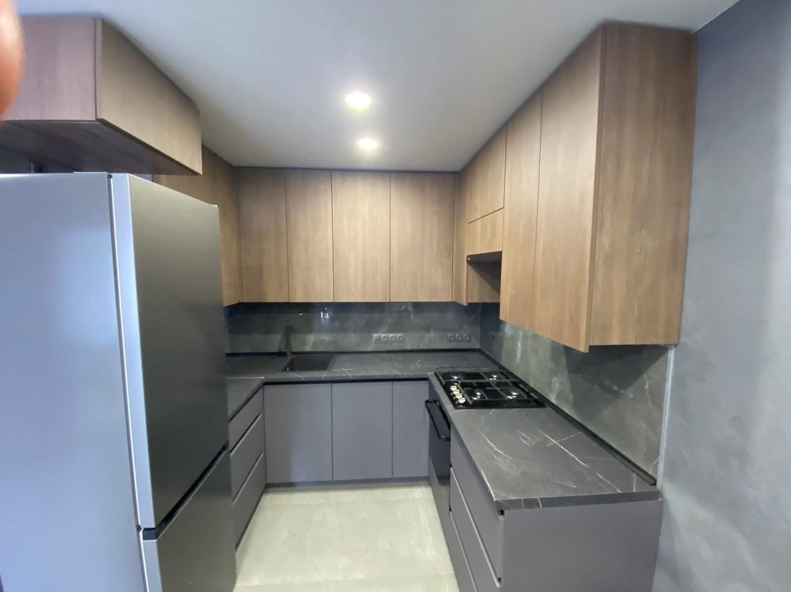 Nowoczesna kuchnia w odcieniach szarości i drewna z marmurowym blatem i sprzętem AGD. Ujęcie z boku, prezentujące funkcjonalność i design mebli na wymiar.