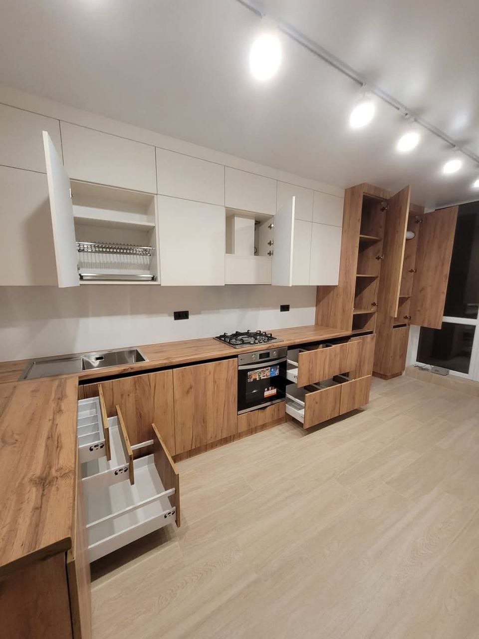 Nowoczesna kuchnia z drewnianymi frontami i białymi szafkami wiszącymi. Otwarta szuflada z organizerem na sztućce, płyta gazowa i piekarnik w zabudowie.