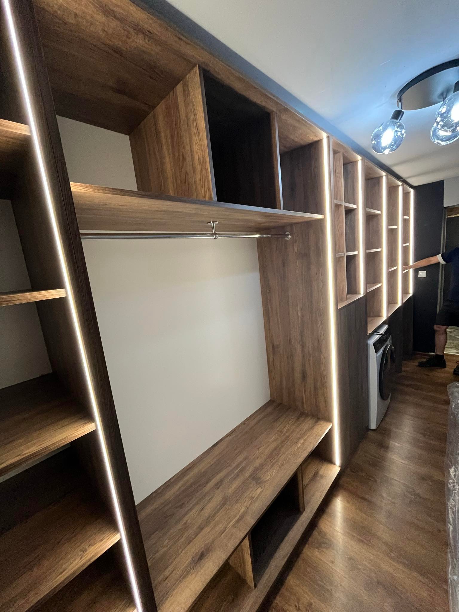 Drewniana szafa wnękowa z oświetleniem LED, wkomponowana pralka. Widoczne półki, drążek na ubrania i schowki. Ujęcie z dołu, podkreślające wysokość zabudowy.