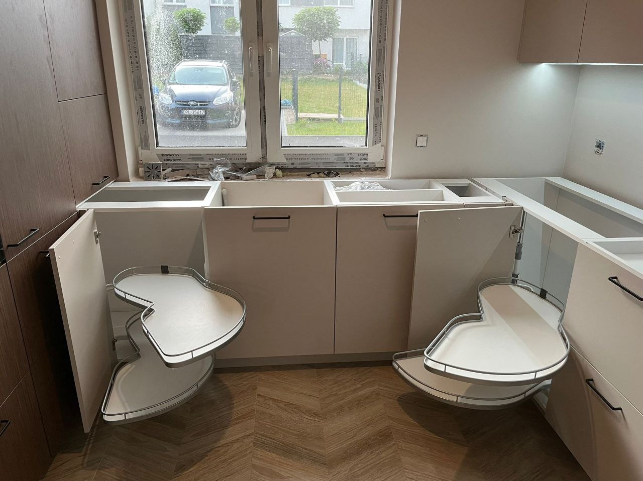 Montaż narożnej szafki kuchennej z wysuwanymi półkami. Widoczne białe fronty, czarne uchwyty i drewniana podłoga. Za oknem zaparkowany samochód.