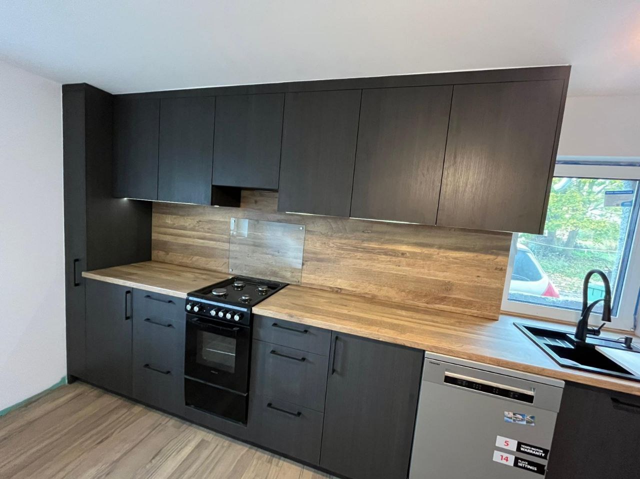 Czarna zabudowa kuchenna z drewnianym blatem i panelem, widok na okno. Nowoczesny design z oświetleniem podszafkowym i sprzętem AGD.