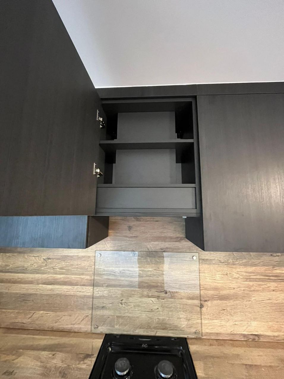 Ciemna, otwarta szafka kuchenna z półkami nad blatem z drewnopodobnym wzorem i szklanym panelem ochronnym, widok z dołu.