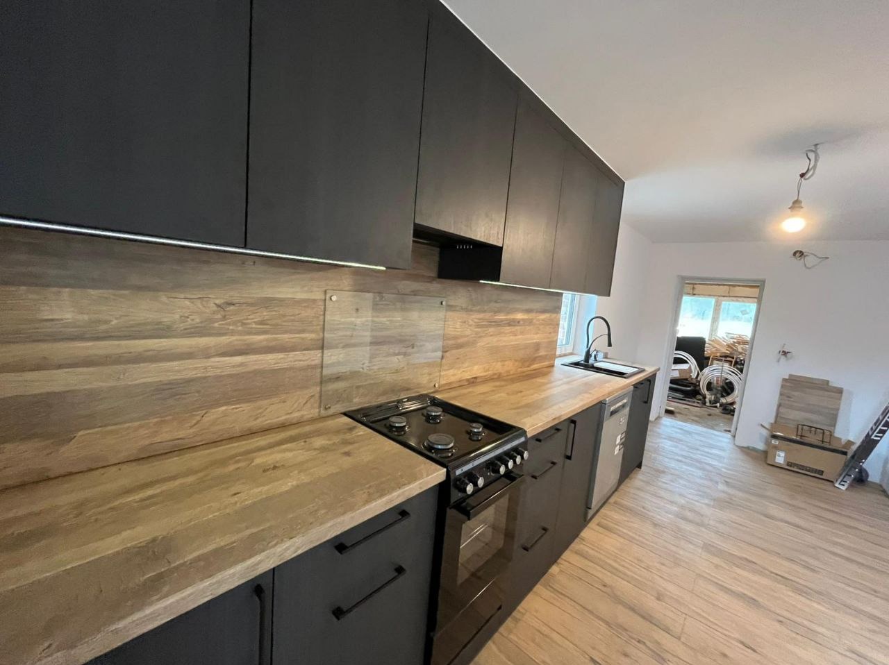 Nowoczesna kuchnia z czarnymi szafkami i drewnianym blatem. Nad blatem podświetlenie LED. W tle widoczne materiały budowlane, sugerujące świeży montaż.