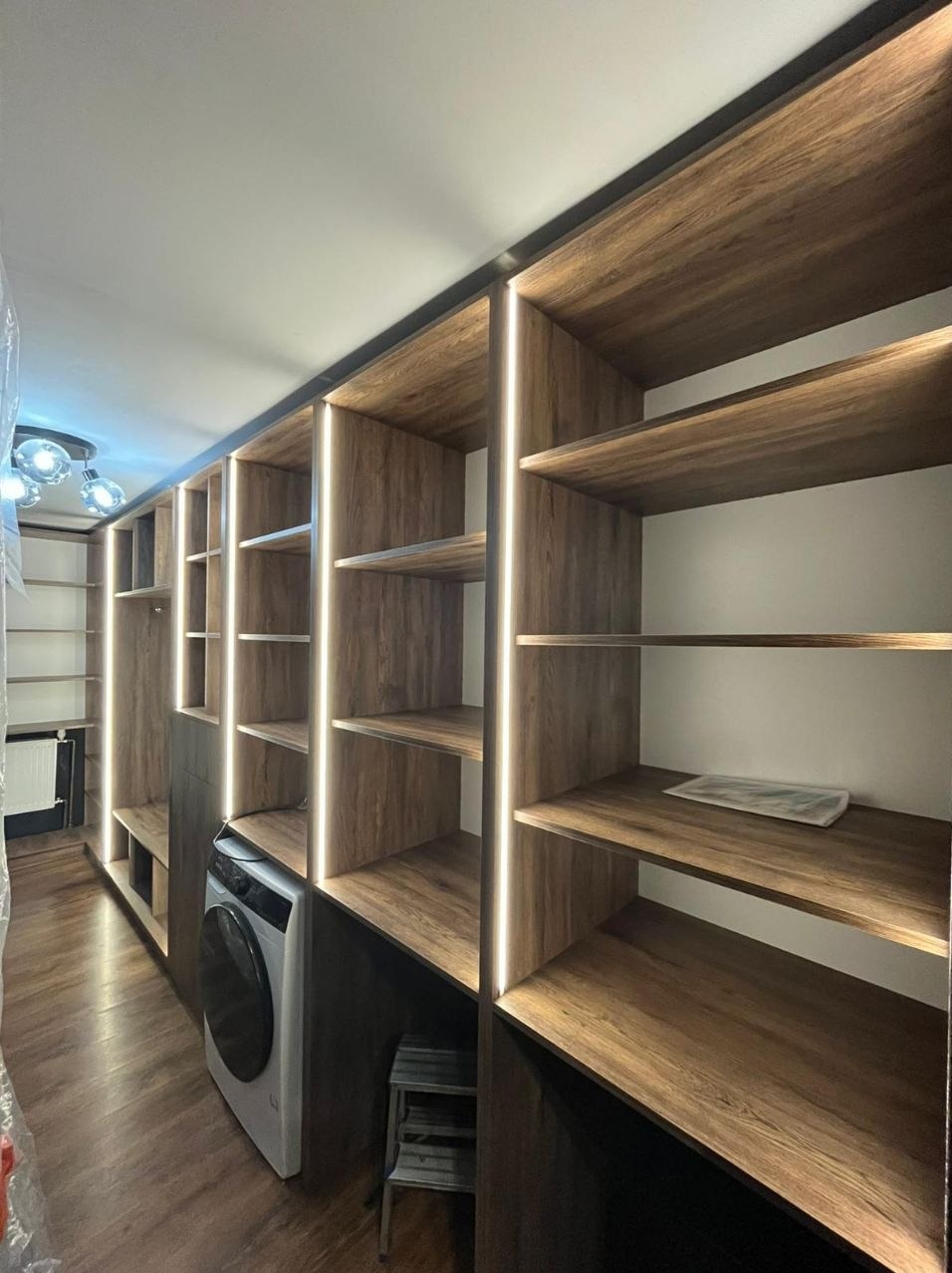 Wnętrze z zabudową drewnianą z podświetleniem LED, integrującą pralkę. Widoczne półki i schowki, tworzące funkcjonalną przestrzeń. Ujęcie z perspektywy podłogi.