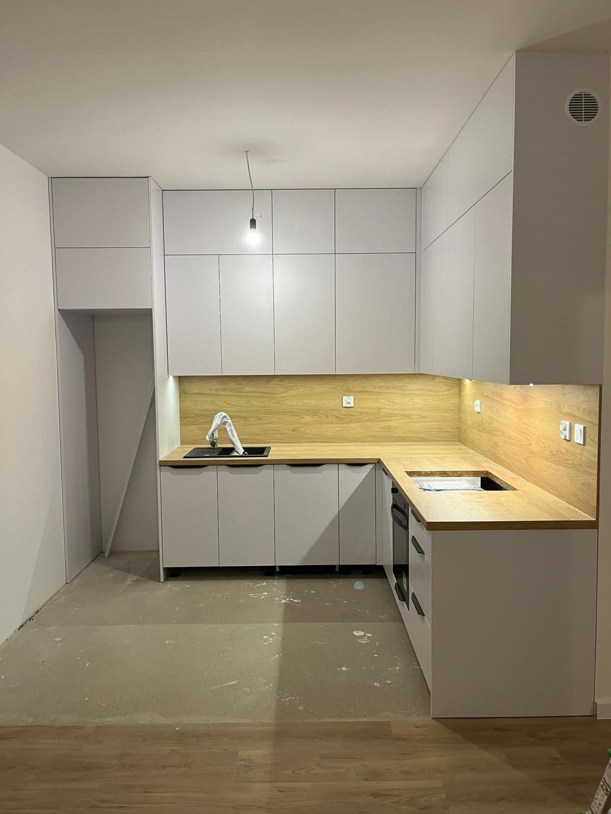Jasna kuchnia z drewnianym blatem i białymi szafkami, z czarnym zlewem i baterią. Widoczne niedokończone wykończenia podłogi i ścian, surowa żarówka.
