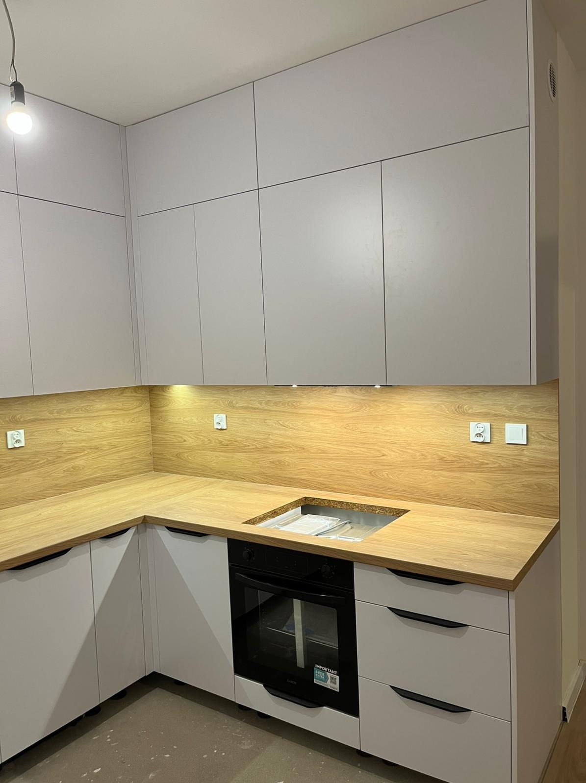 Jasnoszara zabudowa kuchenna z drewnianym blatem i oświetleniem podszafkowym. Widoczny piekarnik i zlewozmywak. Nowoczesny design, minimalistyczne uchwyty.