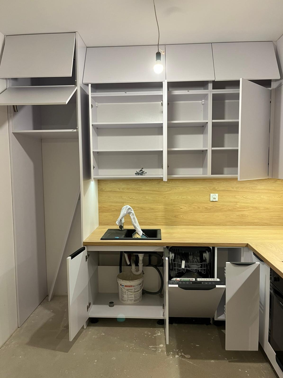 Szara kuchnia z otwartymi szafkami, drewnianym blatem i czarnym zlewem. Widoczna zmywarka i wiadro pod zlewem. Ujęcie z dołu, surowy stan wykończenia.