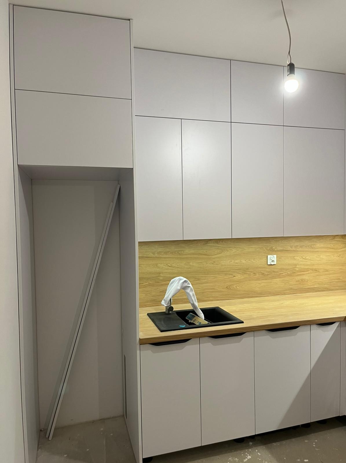 Jasna kuchnia z drewnianym blatem i czarnym zlewem. W szafce widoczna płyta. Nad blatem wisi żarówka. Minimalistyczny design.