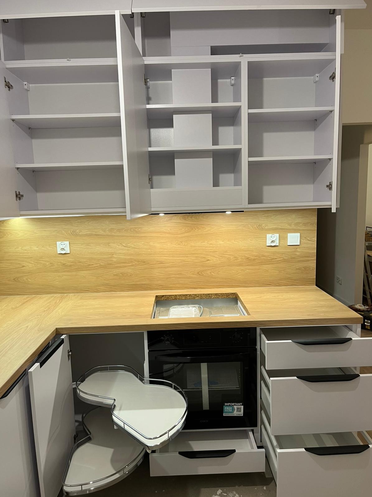 Jasna kuchnia z drewnianym blatem i otwartymi szafkami, widoczny narożny system półek obrotowych i piekarnik. Nowoczesny design z oświetleniem podszafkowym.