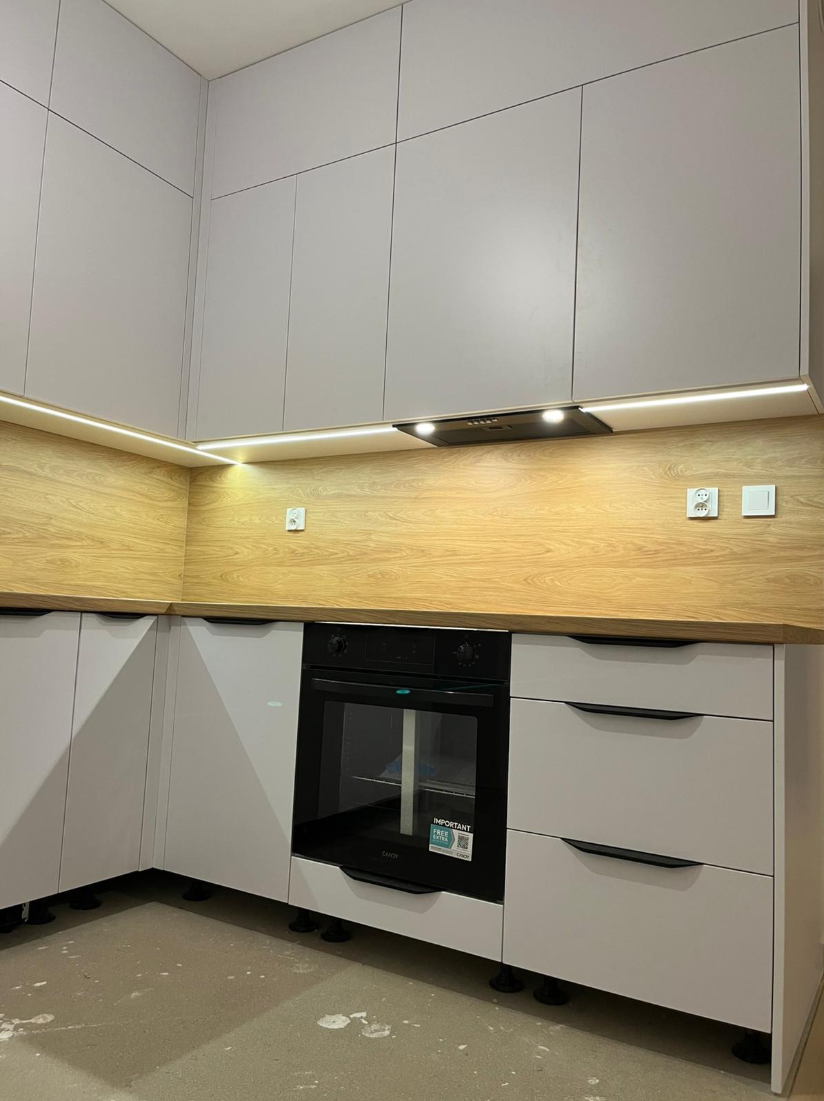 Jasnoszara kuchnia narożna z drewnianym blatem i oświetleniem LED podszafkowym. Widoczny piekarnik i szuflady z czarnymi uchwytami. Nowoczesny design.