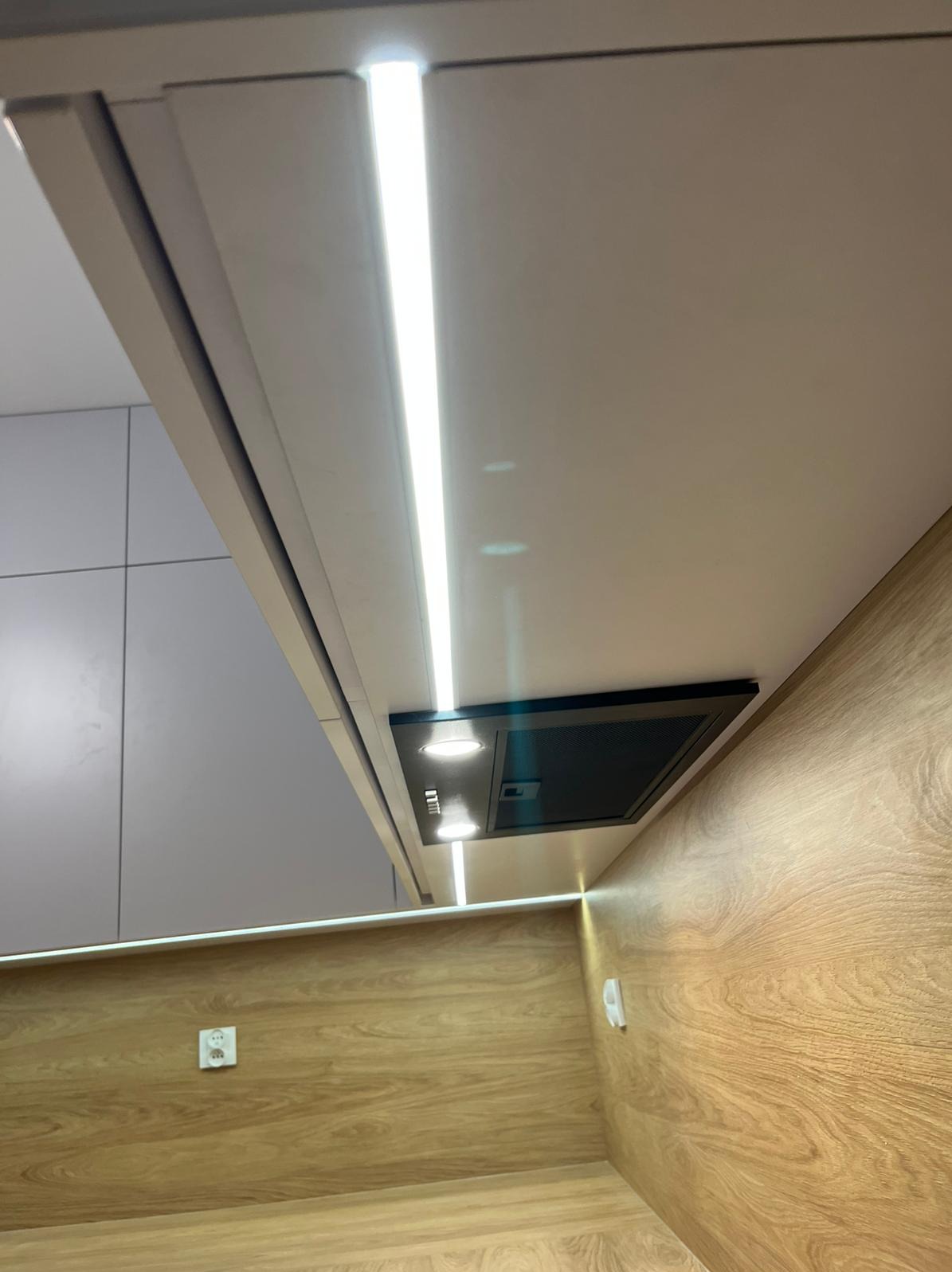 Nowoczesna kuchnia z oświetleniem LED podszafkowym i czarnym okapem, widok z dołu. Jasne fronty szafek i drewnopodobne panele ścienne tworzą spójną całość.