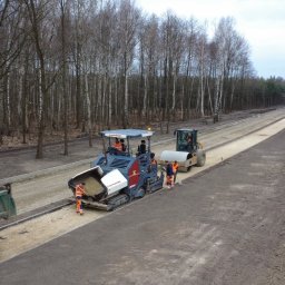 FIL-BUD Sp. z o.o. - Walec i rozkładarka asfaltu przy budowie drogi w lesie, widoczna ciężarówka i pracownicy w pomarańczowych kombinezonach.