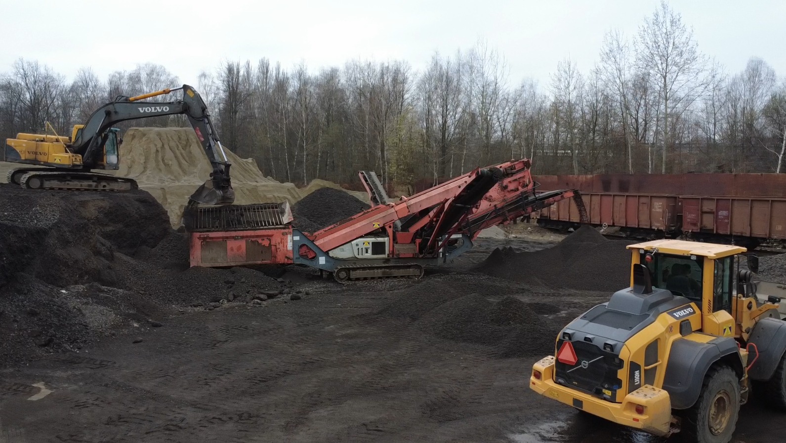 Żółta koparka Volvo nabiera urobek do przesiewacza na gąsienicach, z tyłu wagon kolejowy i sterta piasku.