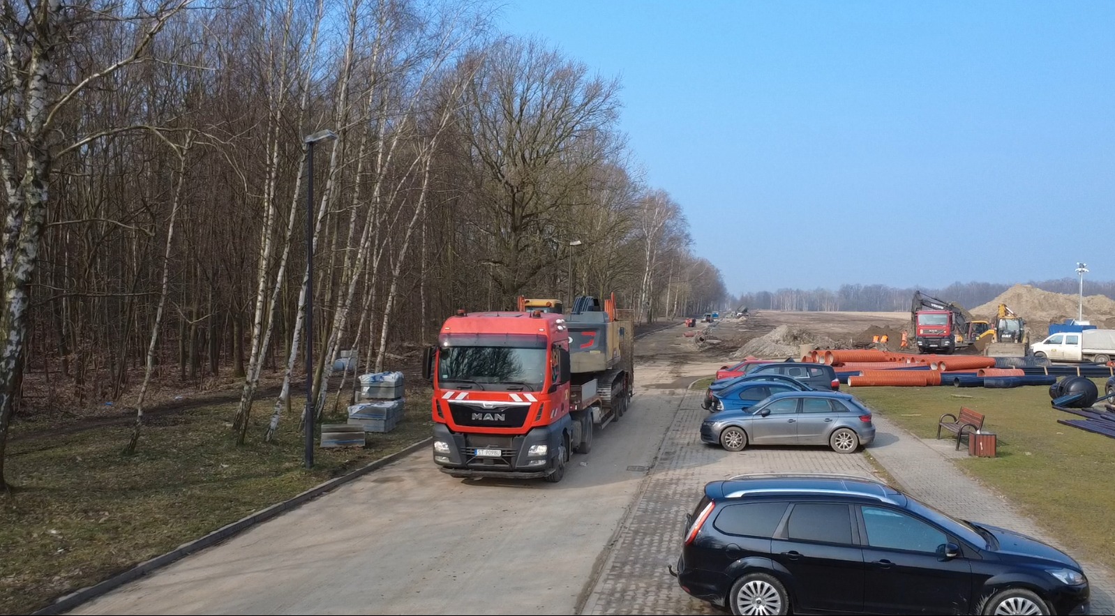 Czerwona ciężarówka MAN przewozi koparkę gąsienicową na naczepie niskopodwoziowej, w tle plac budowy drogi z rurami, koparkami i samochodami osobowymi