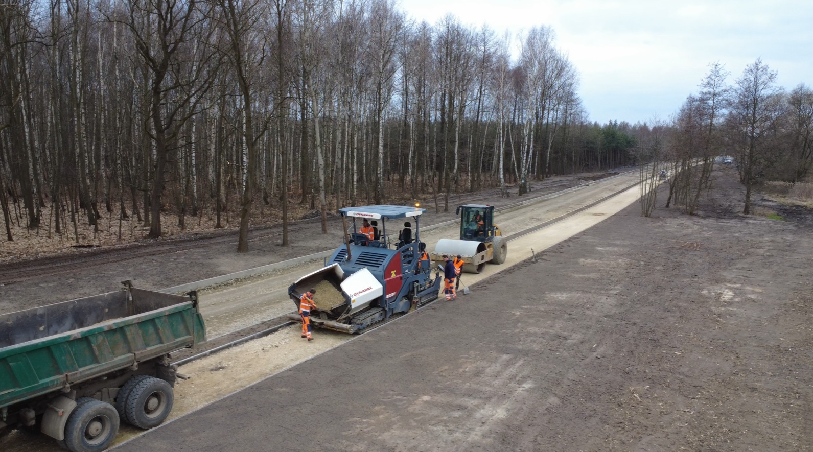 Walec i rozkładarka asfaltu przy budowie drogi w lesie, widoczna ciężarówka i pracownicy w pomarańczowych kombinezonach.