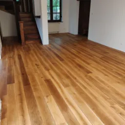 Jasna, odnowiona podłoga drewniana z widocznymi słojami, prowadząca do schodów i okna w przestronnym, jasnym wnętrzu.