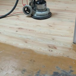 FIRMA USŁUGOWA JACEK HERZBERG - Cyklinowanie drewnianej podłogi w trakcie renowacji, widoczny fragment podłogi przed i po cyklinowaniu, maszyna do cyklinowania z podłączonym odkurzaczem.