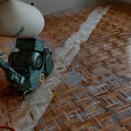 FIRMA USŁUGOWA JACEK HERZBERG - Parkiet w jodełkę częściowo wyszlifowany, widoczna maszyna do szlifowania parkietu marki Hummel i worek na pył.