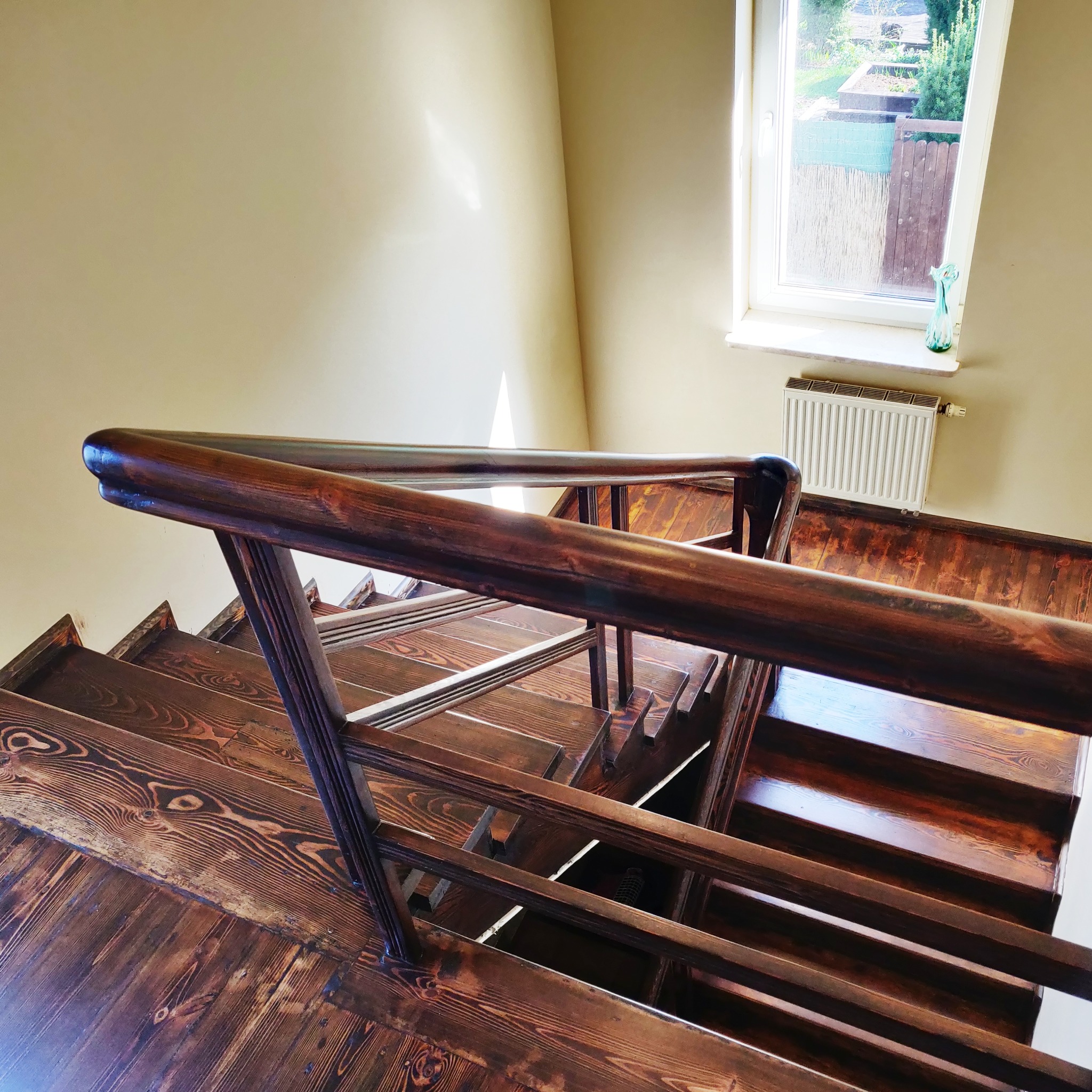Odnowione drewniane schody z ciemnobrązową balustradą, widok z góry, jasne ściany i okno z widokiem na ogród w tle.