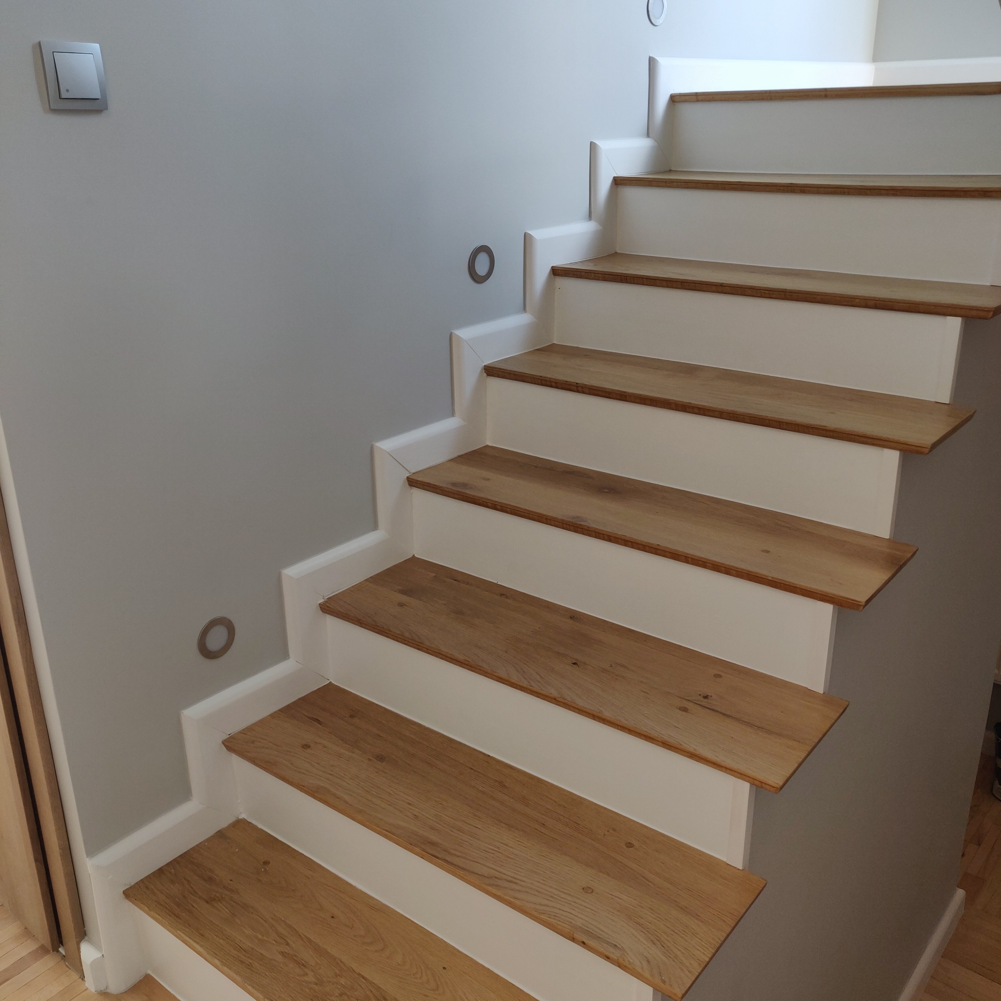 Odnowione drewniane schody z białymi podstopnicami, widoczne oświetlenie punktowe na ścianie i włącznik światła.