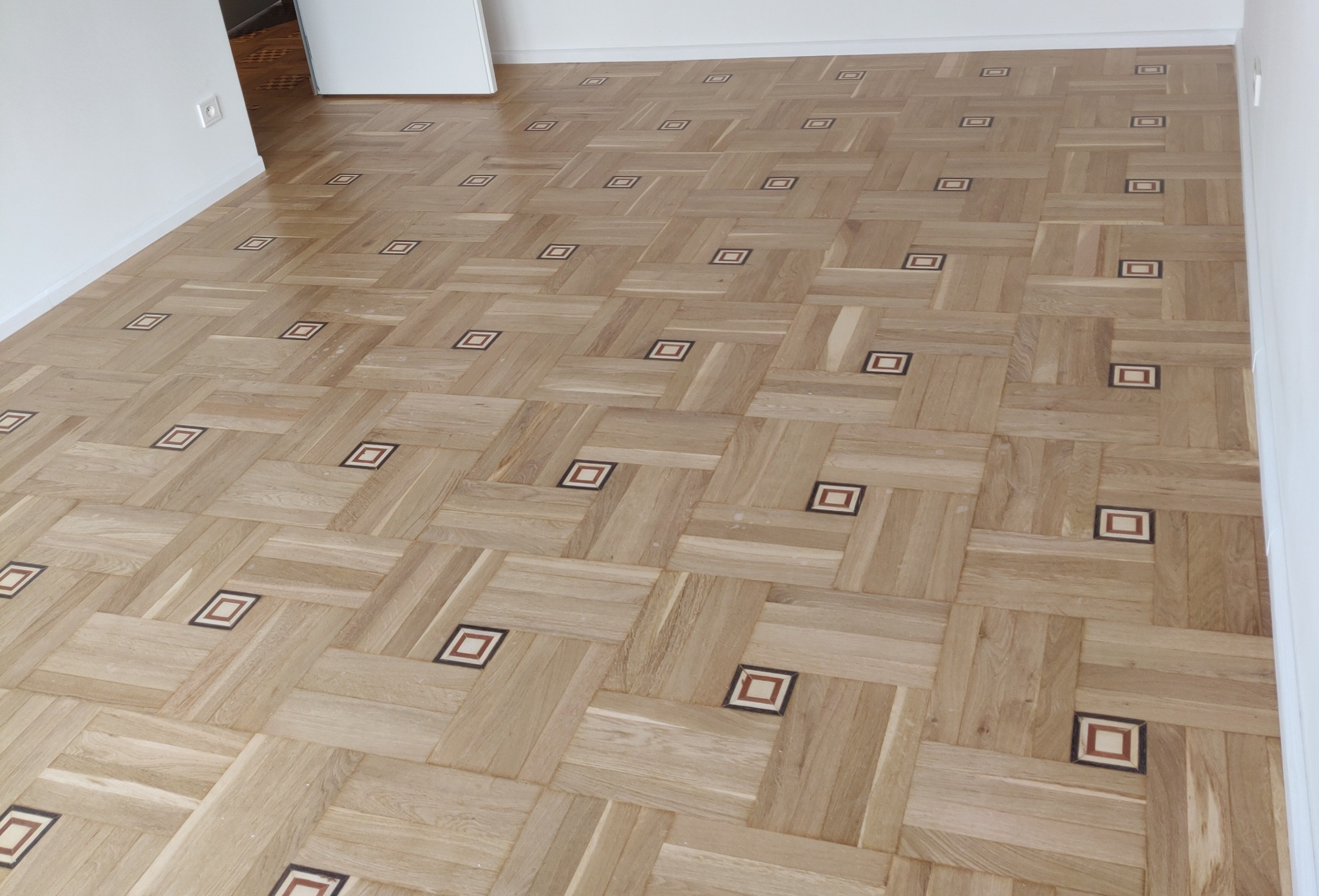 Parkiet dębowy ułożony we wzór szachownicy z ciemniejszymi kwadratowymi intarsjami w każdym polu, świeżo po cyklinowaniu, widoczne delikatne ślady pyłu po obróbce.