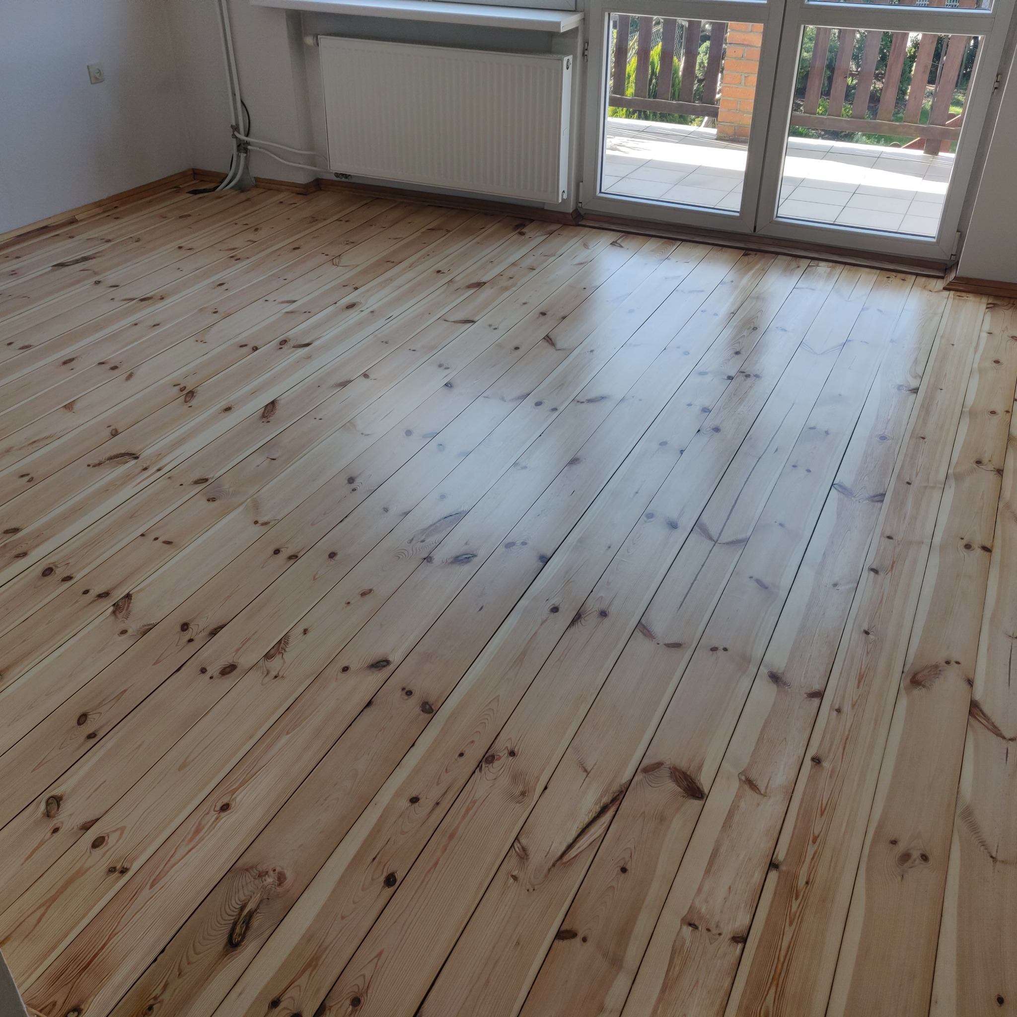 Odnowiona podłoga z jasnego drewna z widocznymi słojami, po zabiegu wygładzania, z oknem balkonowym w tle.