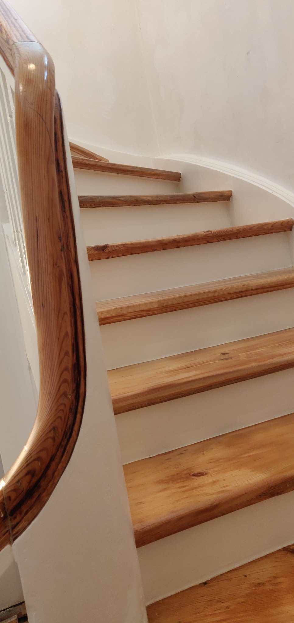 Odnowione drewniane schody z jasnym drewnem i białymi podstopnicami, widok z dołu, z widoczną poręczą o wyraźnym usłojeniu.