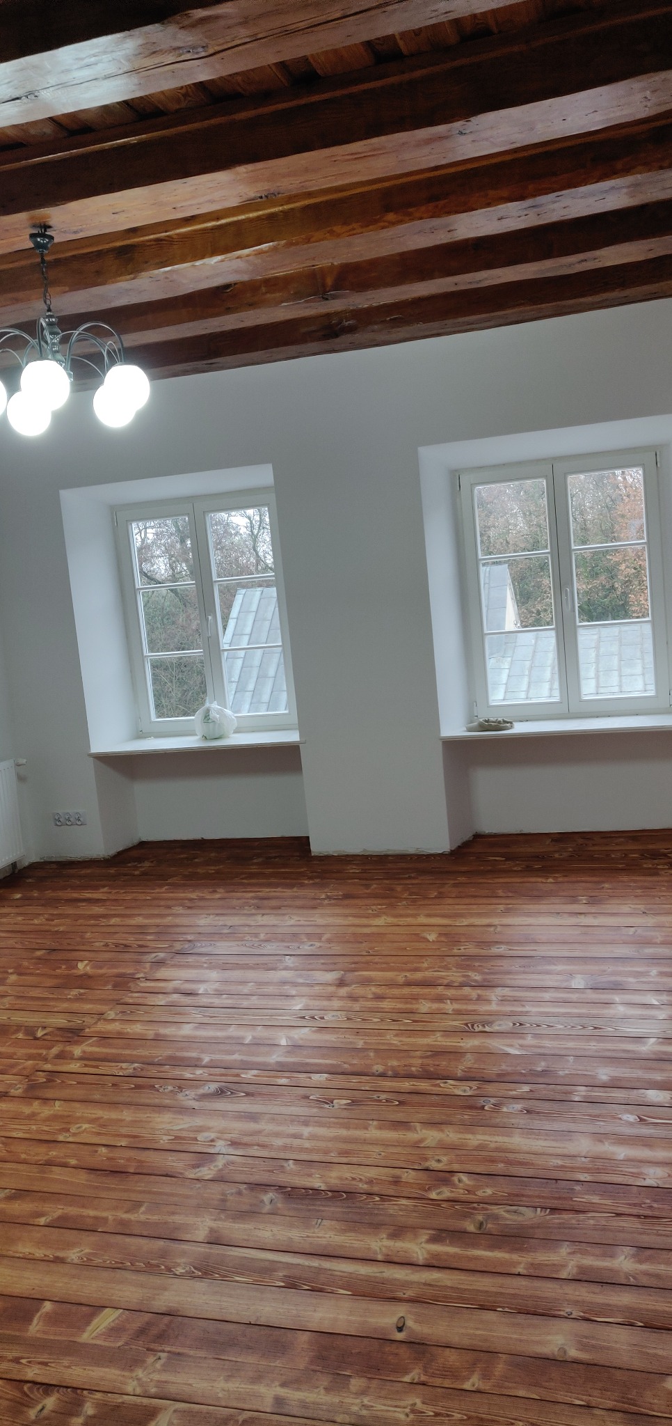 Wycyklinowana podłoga z desek w przestronnym pokoju z dwoma oknami, widoczne drewniane belki stropowe i ozdobny żyrandol.