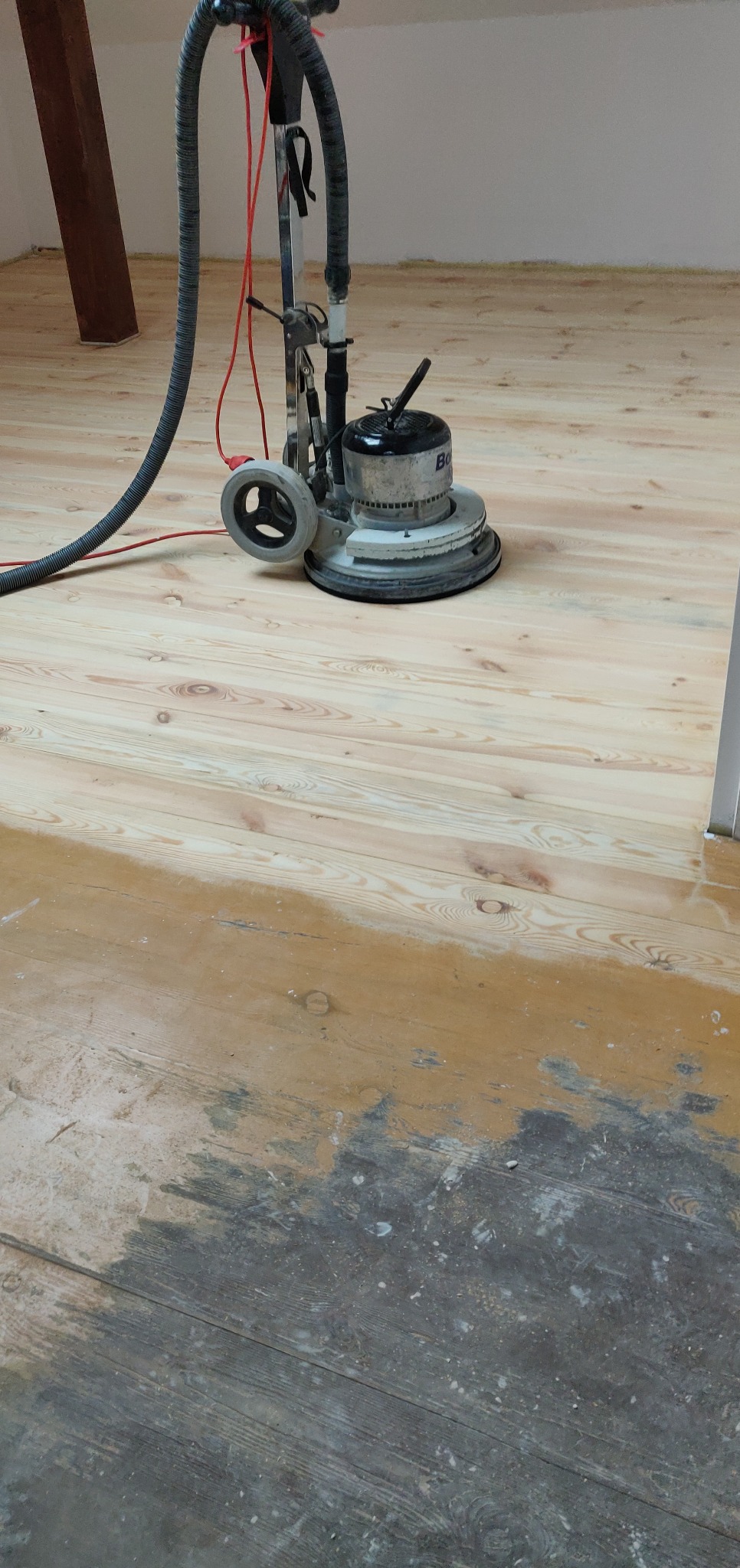 Cyklinowanie drewnianej podłogi w trakcie renowacji, widoczny fragment podłogi przed i po cyklinowaniu, maszyna do cyklinowania z podłączonym odkurzaczem.