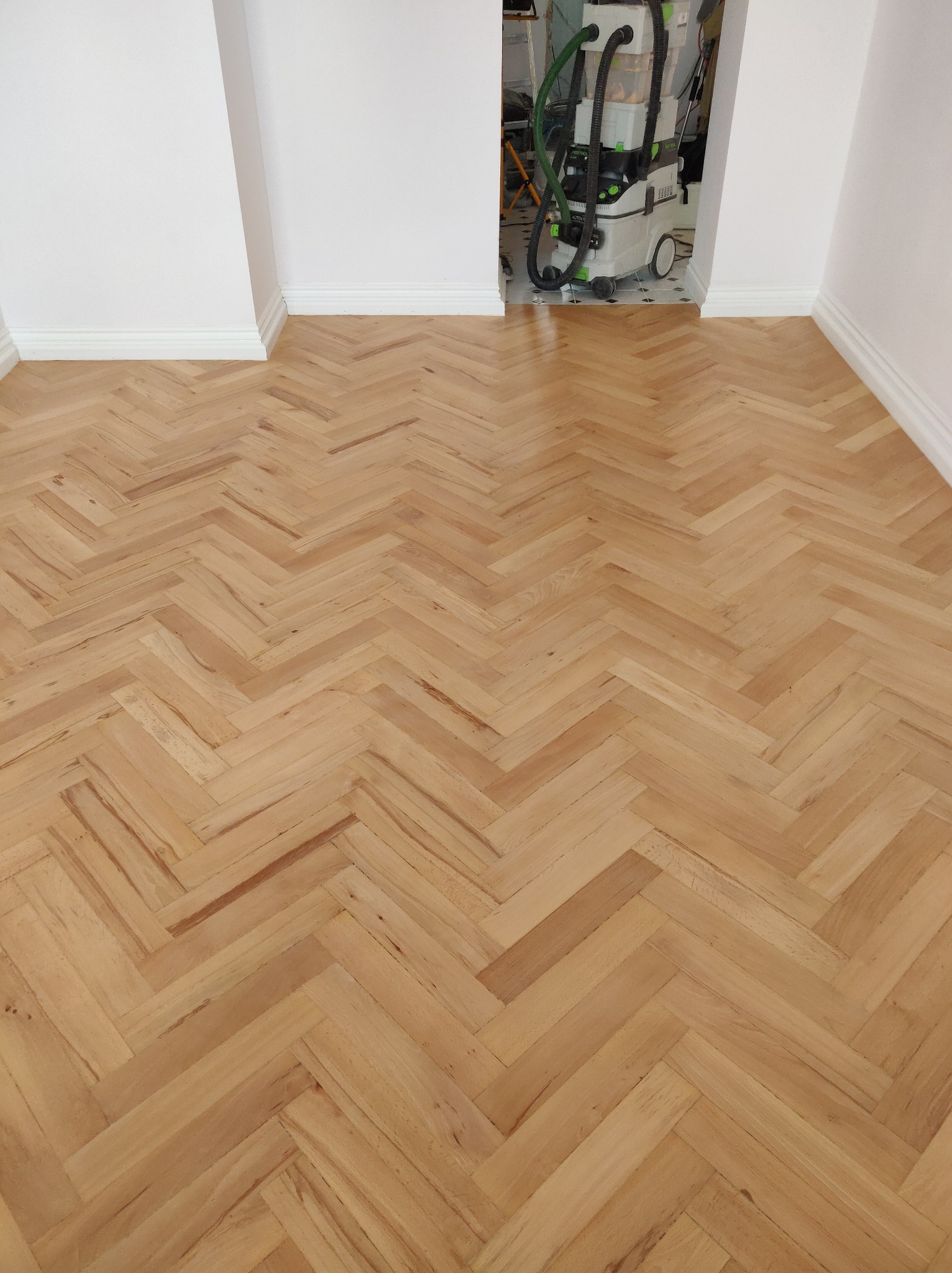 Wycyklinowana podłoga parkietowa ułożona w jodełkę, widoczny odkurzacz przemysłowy w przejściu do innego pomieszczenia.