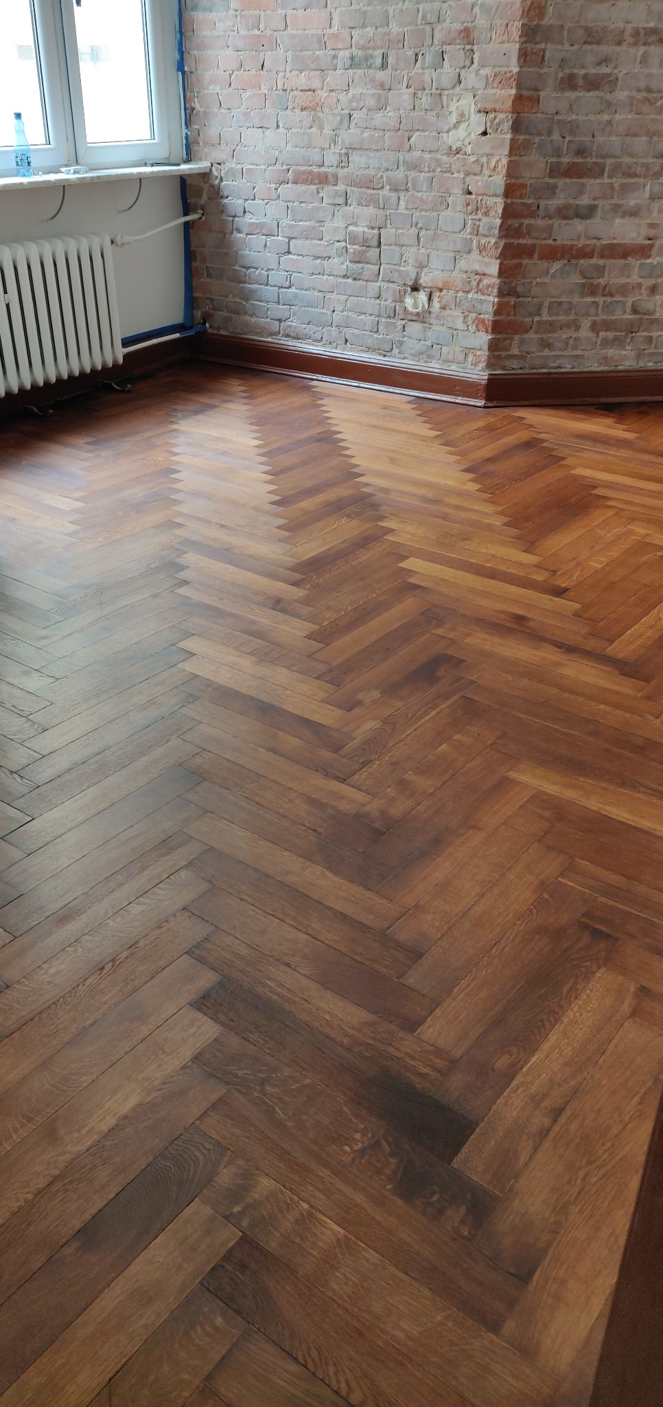 Parkiet ułożony w jodełkę po renowacji, widoczne odnowione drewno i fragment ceglanej ściany w tle.