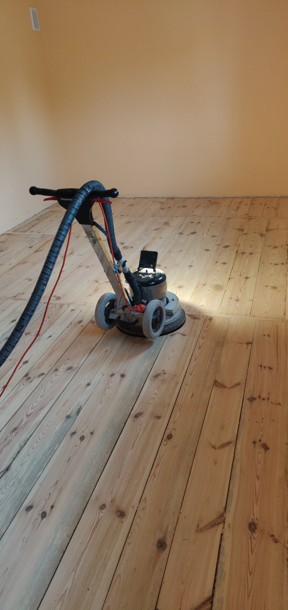 Cyklinowanie drewnianej podłogi deskowej maszyną z odkurzaczem; widoczny efekt szlifowania na jasnym drewnie z wyraźnym usłojeniem.