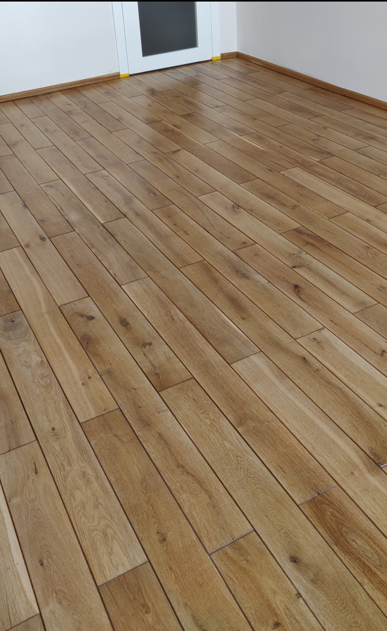 Wycyklinowana podłoga z drewna o jasnobrązowym odcieniu, ułożona w jodełkę, widoczna przy białej ścianie i drzwiach z szybą.