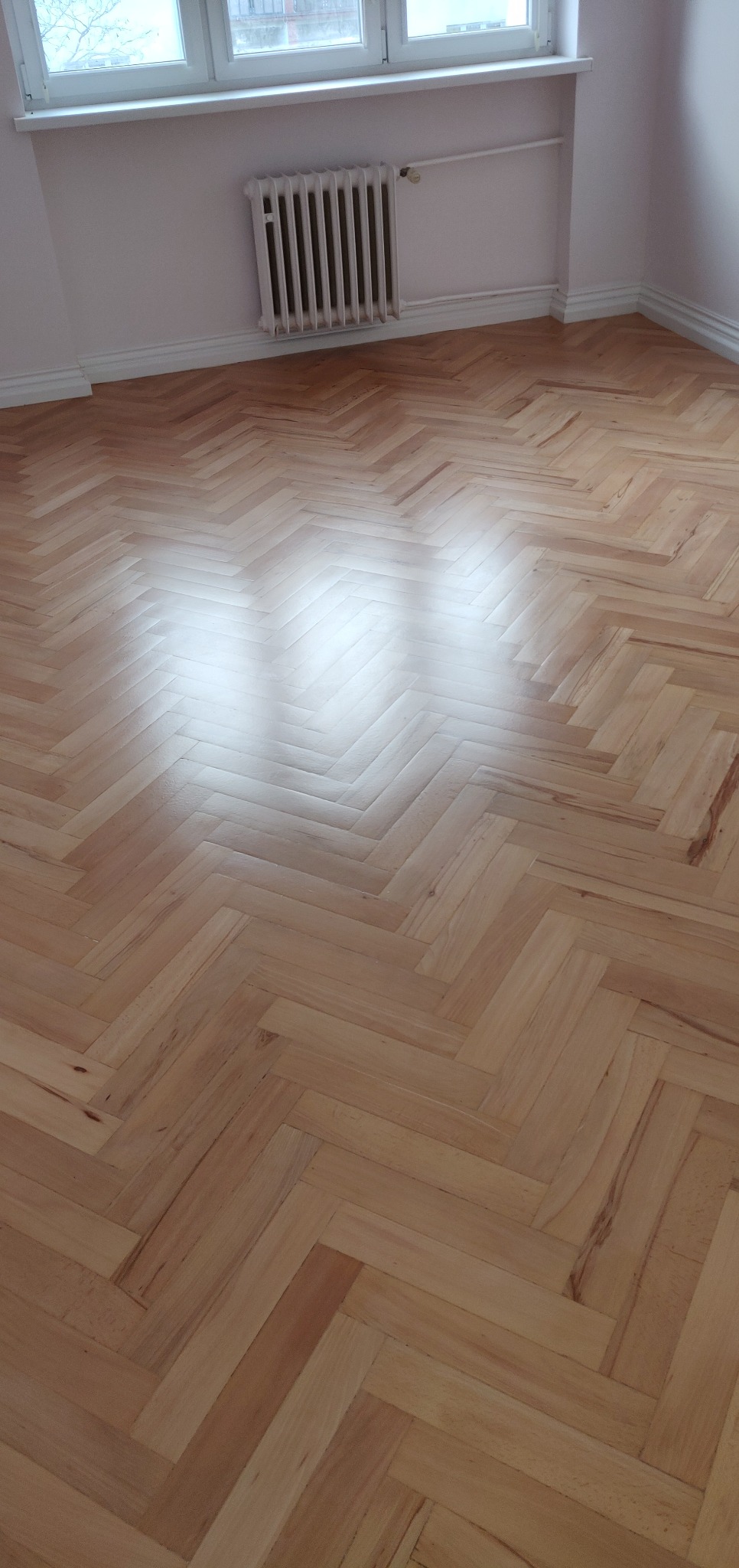 Parkiet ułożony w jodełkę po renowacji, jasne drewno, widoczny grzejnik i fragment okna.